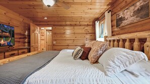 A wooden cabin bedroom with a bed, pillows, a television, and a ceiling fan.