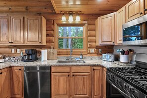 Una cocina con armarios de madera, lavavajillas de acero inoxidable, estufa a gas y ventana con vistas a árboles.