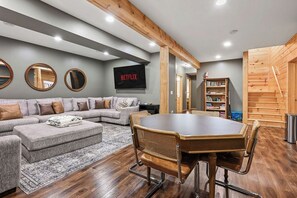 A living room with a grey sofa, a wooden dining table with chairs, a flat-screen TV, and a staircase. - Beautiful 5-bedroom lakefront cabin with AC in charming Necedah! (Necedah)