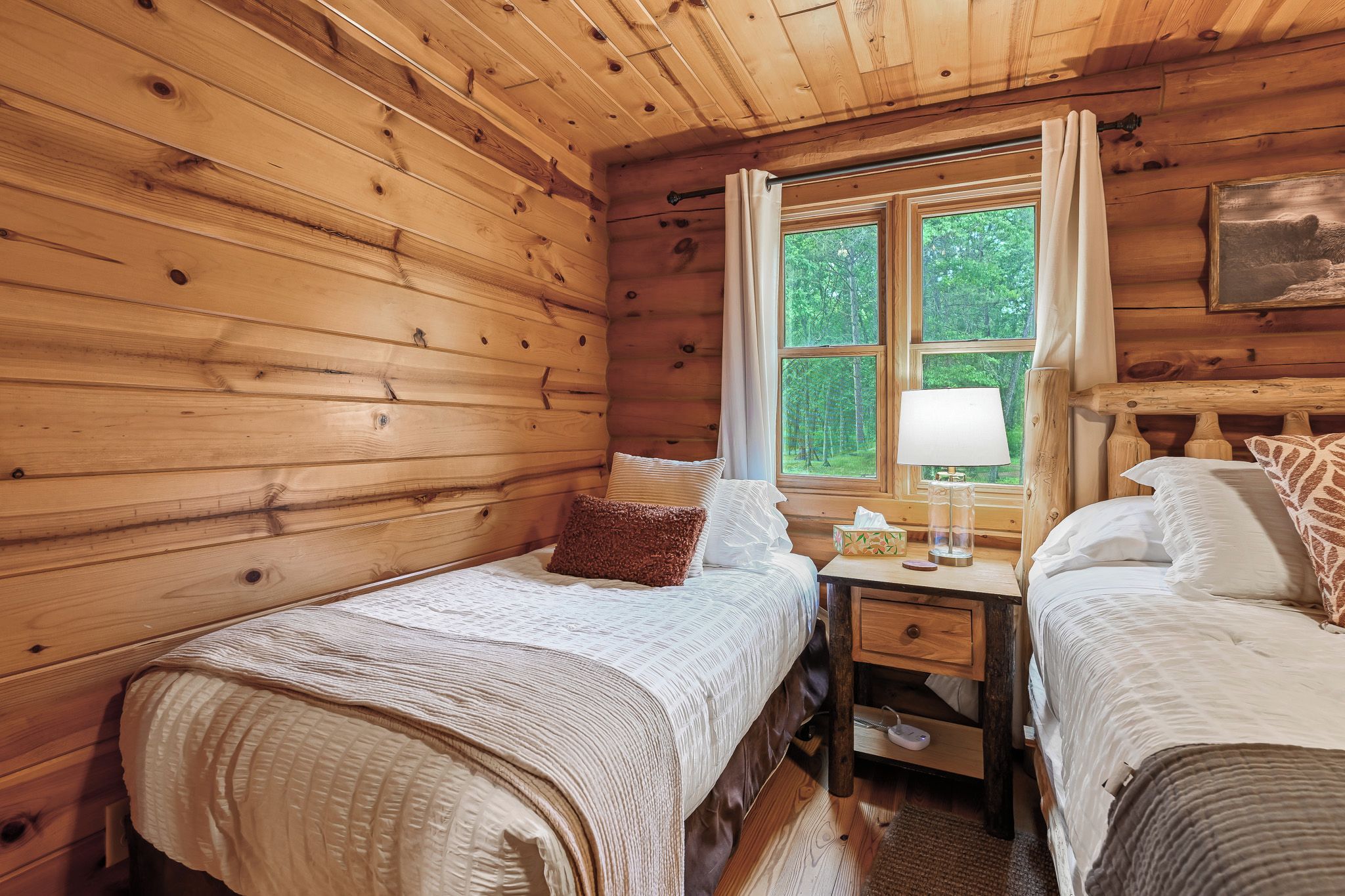 A cabin bedroom with two beds, a nightstand, a window with curtains, and a painting on the wall.