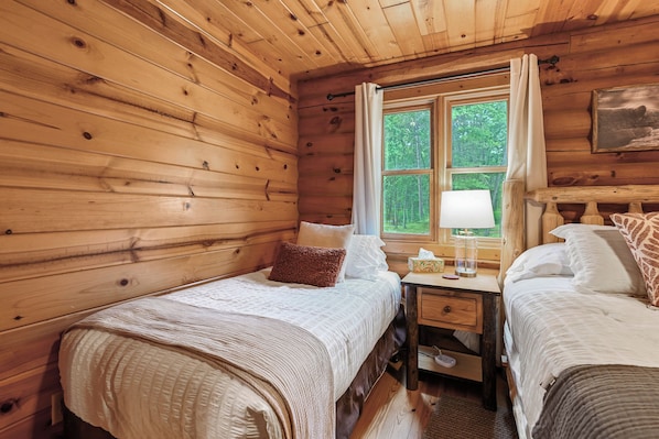 Un dormitorio de cabaña con dos camas, una mesita de noche, una ventana con cortinas y un cuadro en la pared.