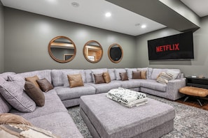 Een moderne woonkamer met een grijze hoekbank, drie ronde spiegels, een Netflix-tv en een gebogen ottoman met een patroonkleed.