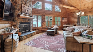 L’intérieur d’une cabane en rondins, avec une cheminée en pierre, un plancher en bois et une grande fenêtre donnant sur un plan d’eau.
