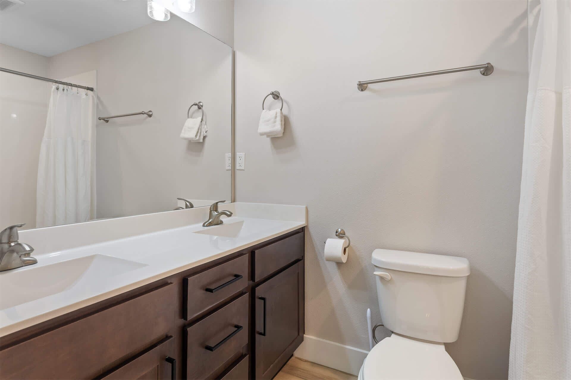 A bathroom with a large mirror, two sinks, a toilet, and a towel rack.