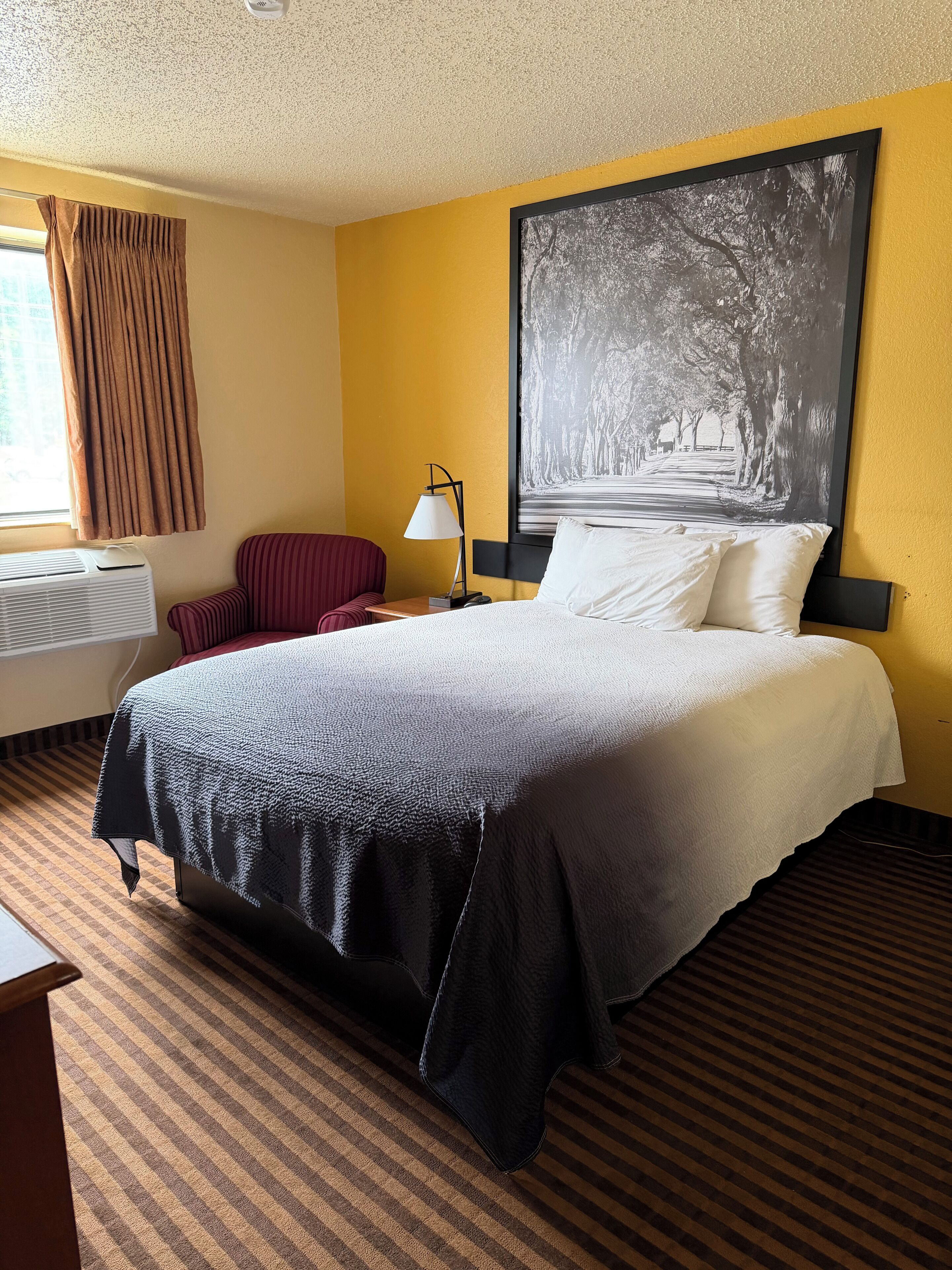 A hotel room with a bed, a chair, a desk, a lamp, and a large framed picture on the wall.