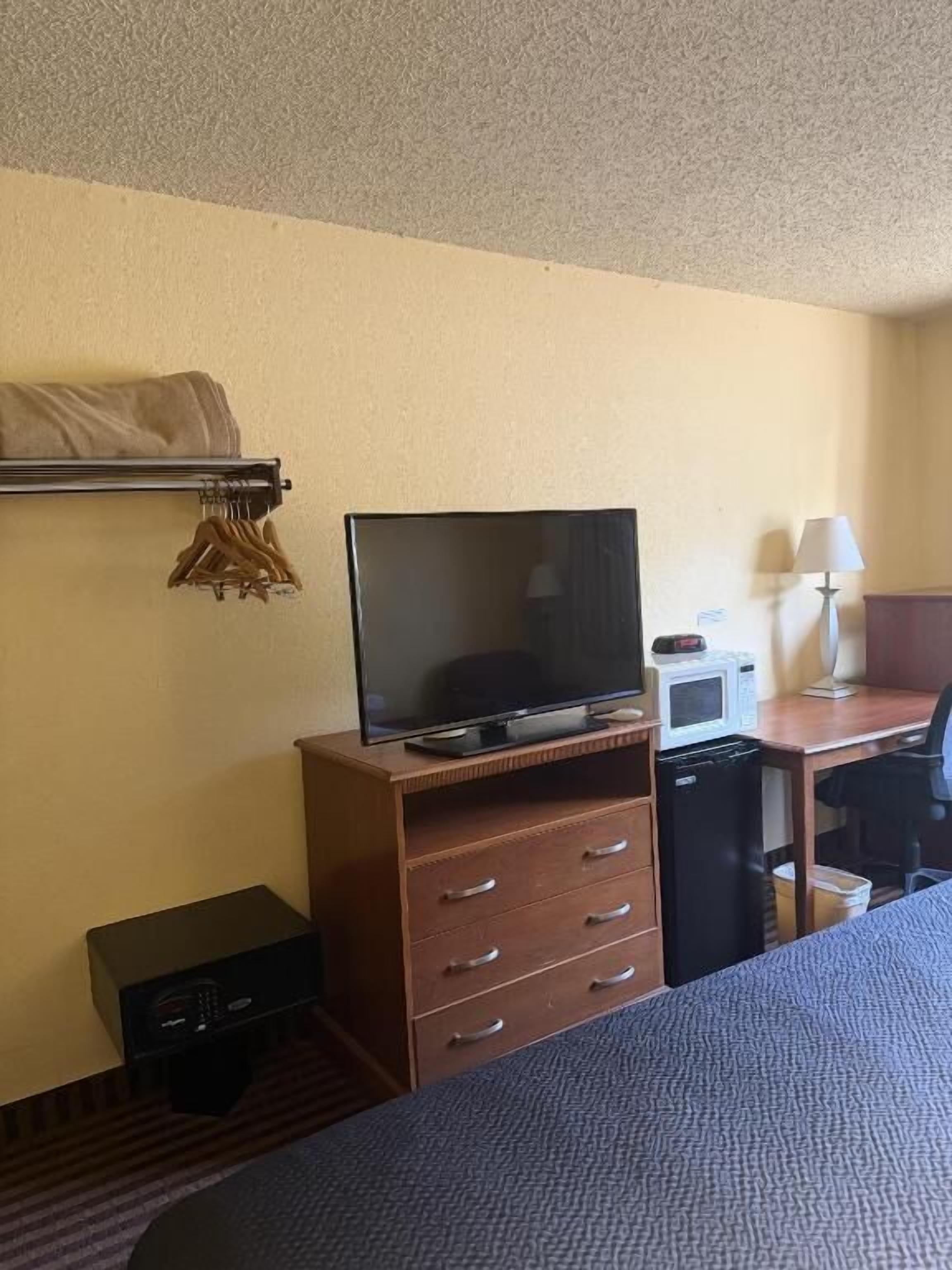A hotel room with a bed, a TV on a wooden stand, a microwave, a small desk, and a lamp.