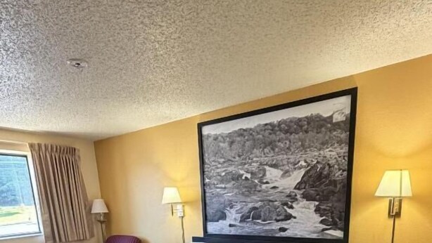 Une chambre d’hôtel avec un grand lit, deux lampes de chevet, une chaise, une fenêtre avec des rideaux et un tableau représentant un paysage, encadré, accroché au mur.