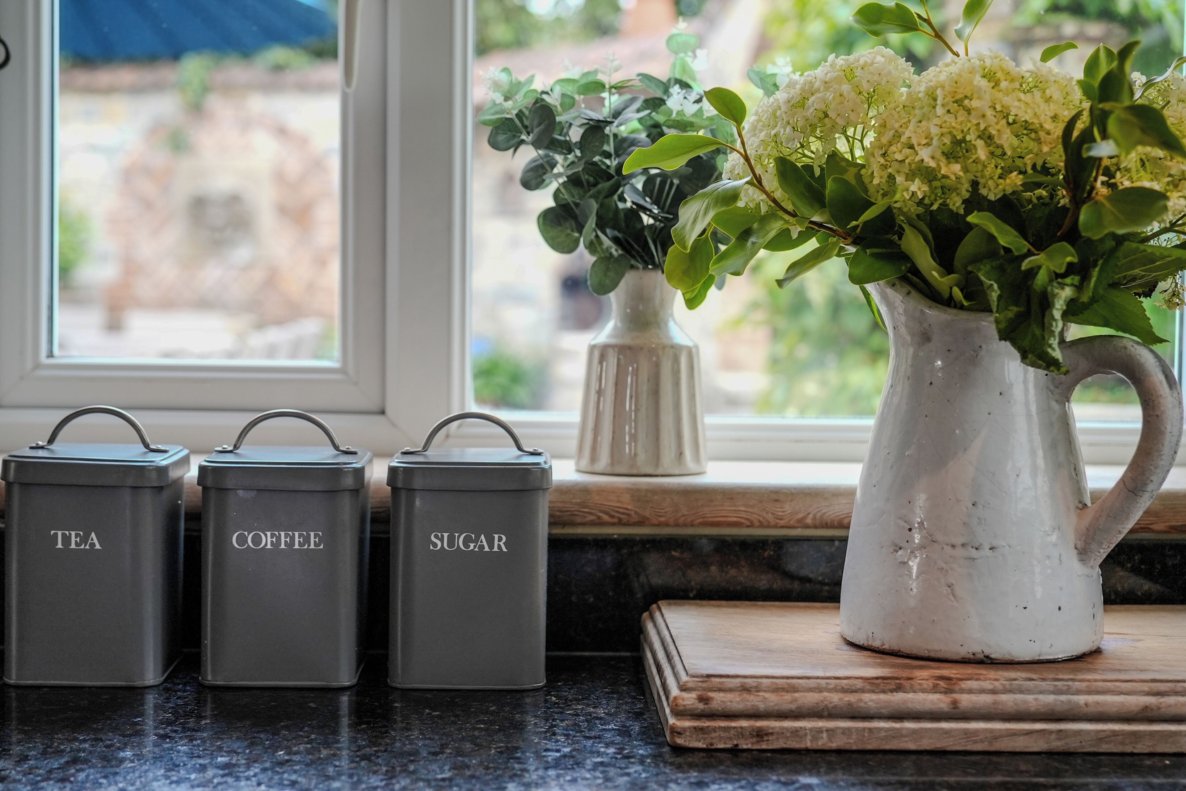 Una encimera de cocina con tres recipientes grises etiquetados 'TEA', 'COFFEE' y 'SUGAR', y una jarrón blanco con flores.