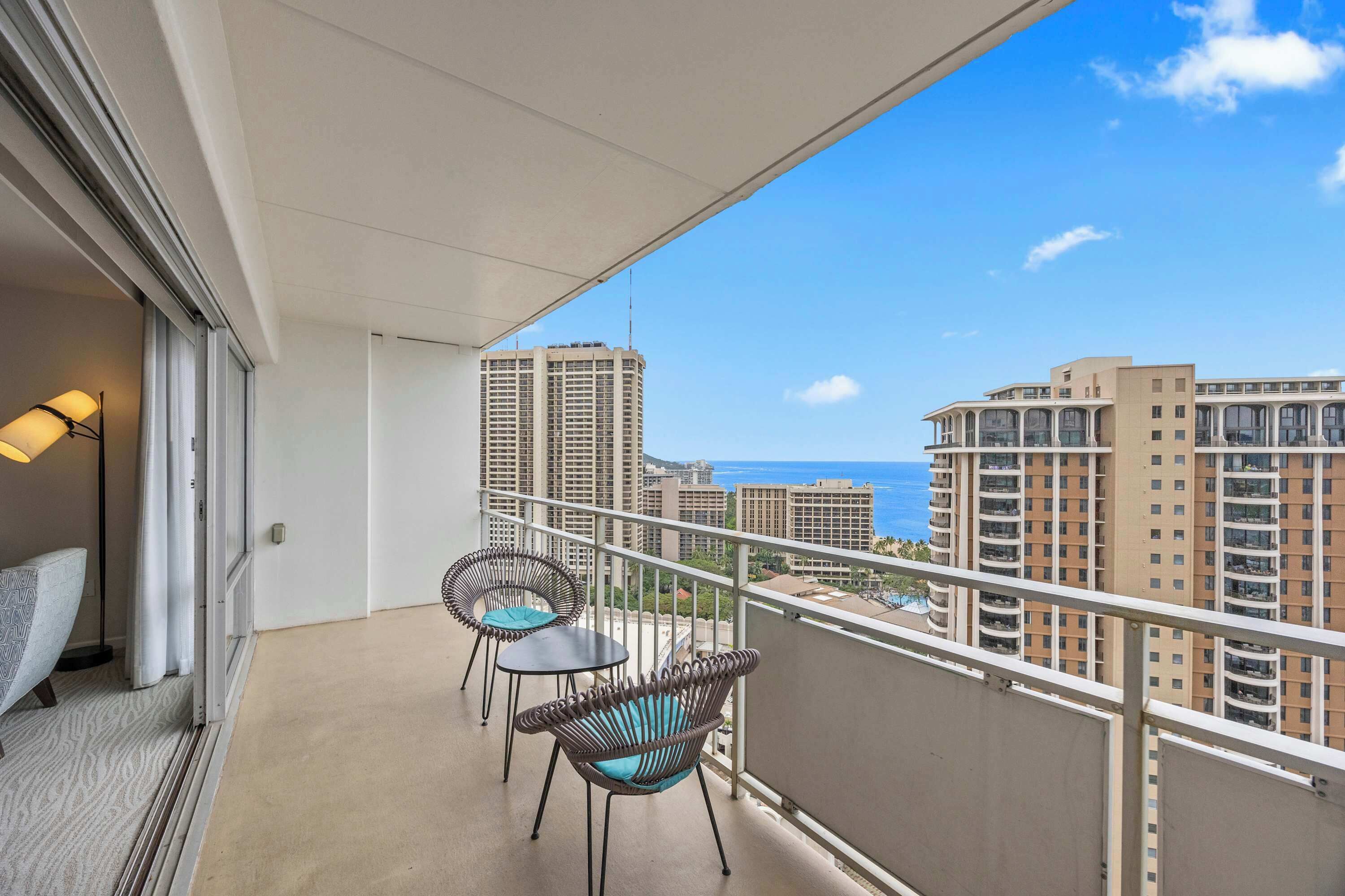 A balcony with two wicker chairs and a small table, overlooking a cityscape with high-rise buildings and a view of the ocean.