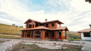 A two-story wooden house with a balcony, surrounded by a gravel driveway and a hilly landscape.