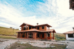 Una casa de madera de dos pisos con balcón, rodeada por un camino de grava y un paisaje montañoso.