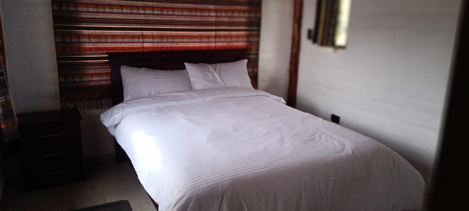 Un lit bien fait dans une chambre d’hôtel, avec une tête de lit à rayures, une table de chevet et un luminaire fixé au mur.