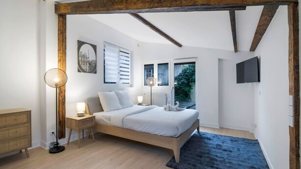 Ein Schlafzimmer mit einem Bett, Nachttischlampen, einer Kommode, einer Stehlampe, einem Fernseher und Blick auf Grünfläche.