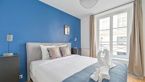 Una habitación de hotel con cama, mesita de noche, lámpara, ventana con cortinas y vistas a edificios.
