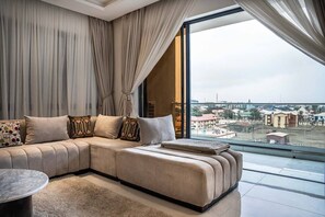 Een moderne woonkamer met een ruime hoekbank, een ronde salontafel en uitzicht op de stad door grote ramen.