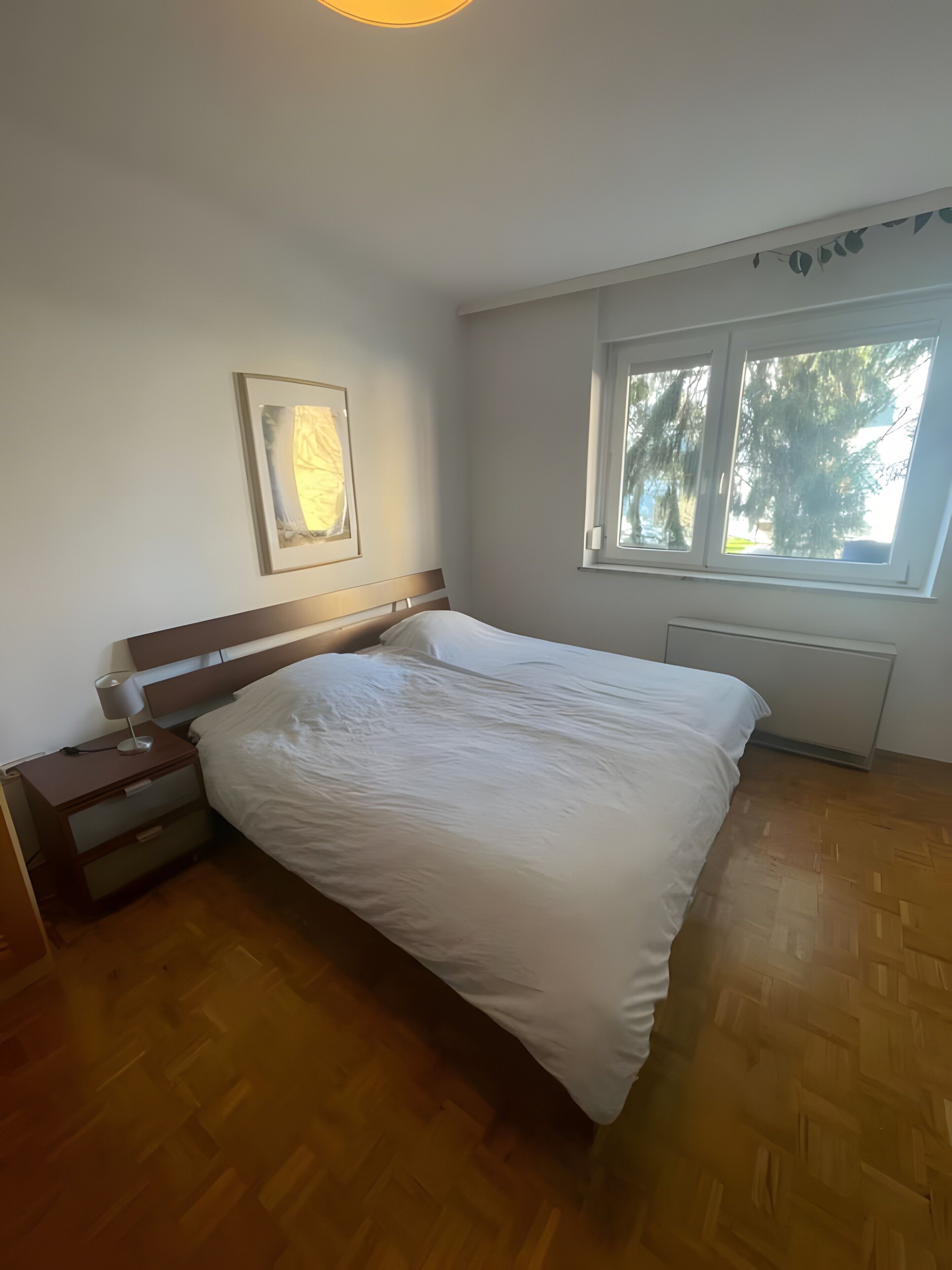 Ett sovrum med en säng, ett sängbord, en lampa, en inramad tavla, ett fönster och ett element.