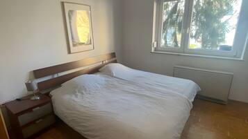 Ett sovrum med en säng, ett sängbord, en lampa, en inramad tavla, ett fönster och ett element.