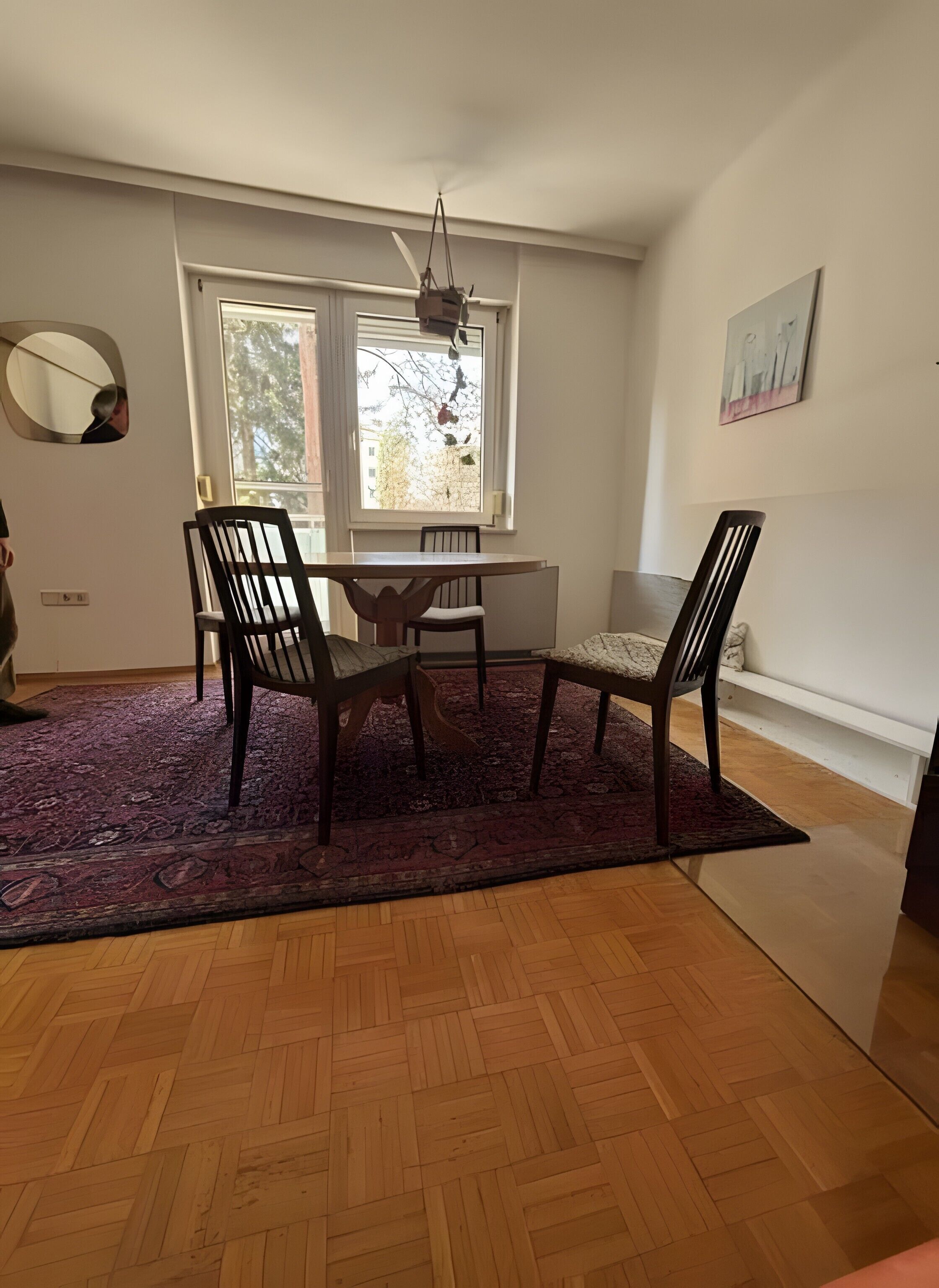 Un coin repas avec une table et des chaises, une fenêtre donnant sur des arbres, un ventilateur de plafond et un miroir fixé au mur.