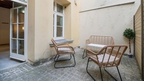 En lille udendørs siddeplads med fletstole og et glasbord, ved siden af en bygning med et vindue og en dør.