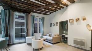 En moderne stue med en sofa, to lænestole, et spisebord og et skrivebord. Der er vinduer med gardiner, et loft med synlige bjælker og en gulvlampe.