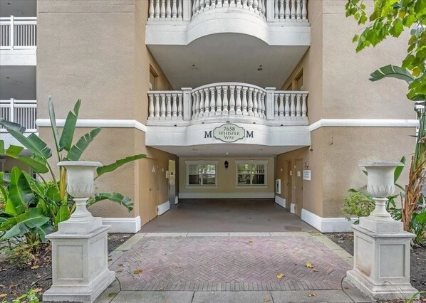 Entrada de un edificio con un balcón doble, un letrero que dice '7658 WHISPER WAY' y dos macetas grandes.