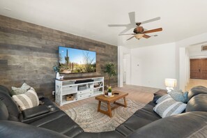 A modern living room with a flat-screen TV, a ceiling fan, a wooden entertainment unit, and a large rug. - Spacious 4-Bedroom Home ~ Backyard BBQ Oasis (Phoenix)