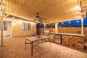 An outdoor patio with a table, a ceiling fan, string lights, a brick wall, and a stone countertop. - Spacious 4-Bedroom Home ~ Backyard BBQ Oasis (Phoenix)