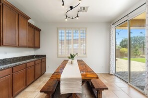 A kitchen with wooden cabinets, a granite countertop, a wooden dining table with benches, a white table runner, a potted plant, and a large window with blinds. - Spacious 4-Bedroom Home ~ Backyard BBQ Oasis (Phoenix)