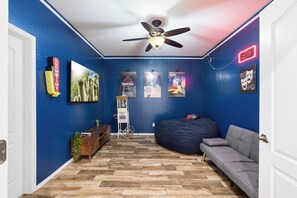Une pièce aux murs bleus, avec un ventilateur au plafond, un canapé gris, un fauteuil poire, un plancher en bois et des affiches de films.