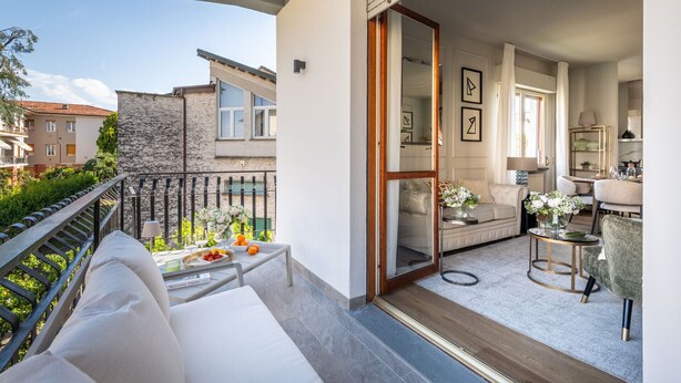 En balkon med en sofa, et spisebord med frugt og udsigt til omkringliggende bygninger.