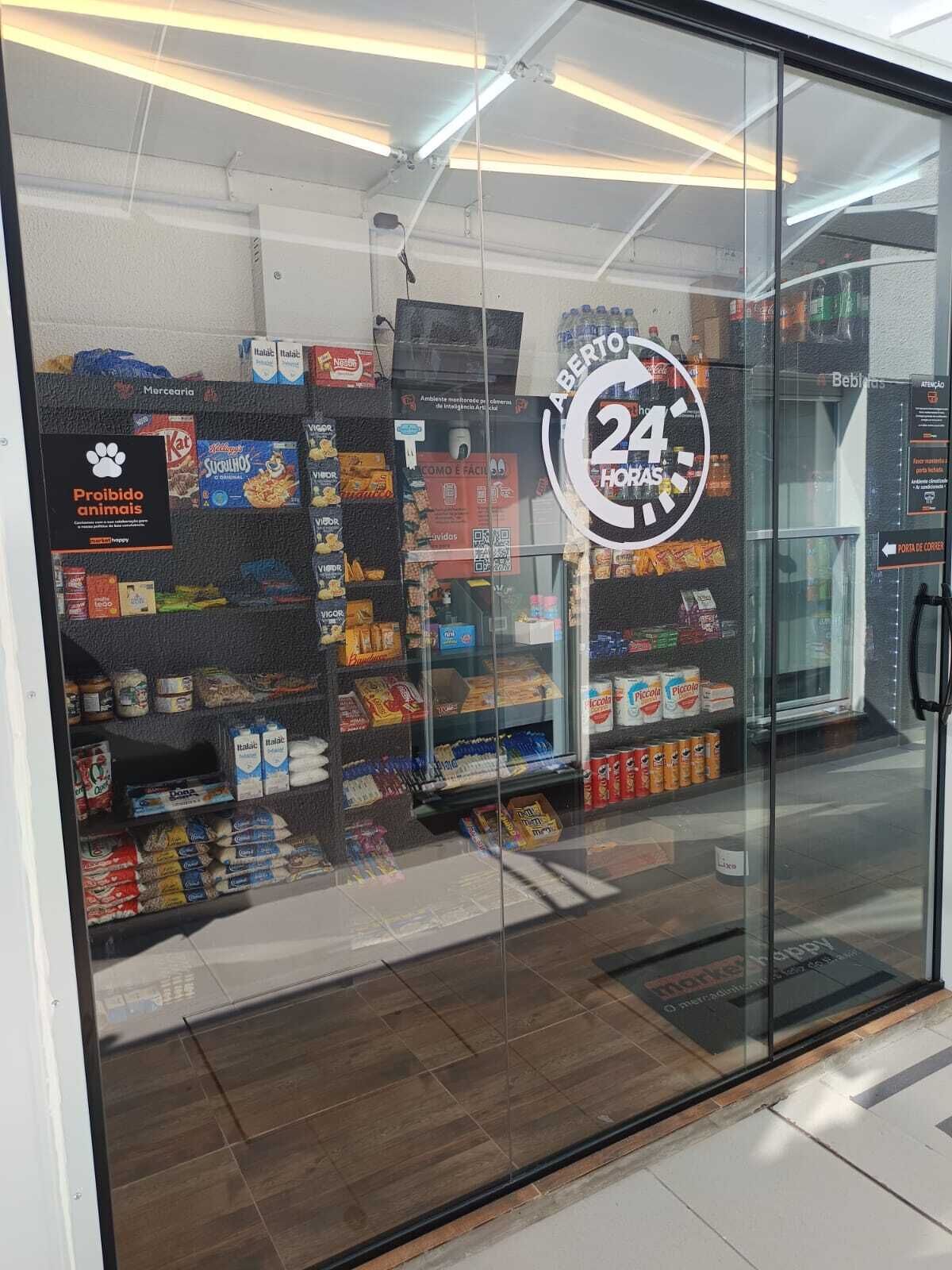 Loja com placa indicando 24 horas, prateleiras cheias de lanches e um aviso de 'Proibido animais'.