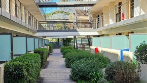 A corridor with a wooden deck, greenery, and a multi-level building with balconies.