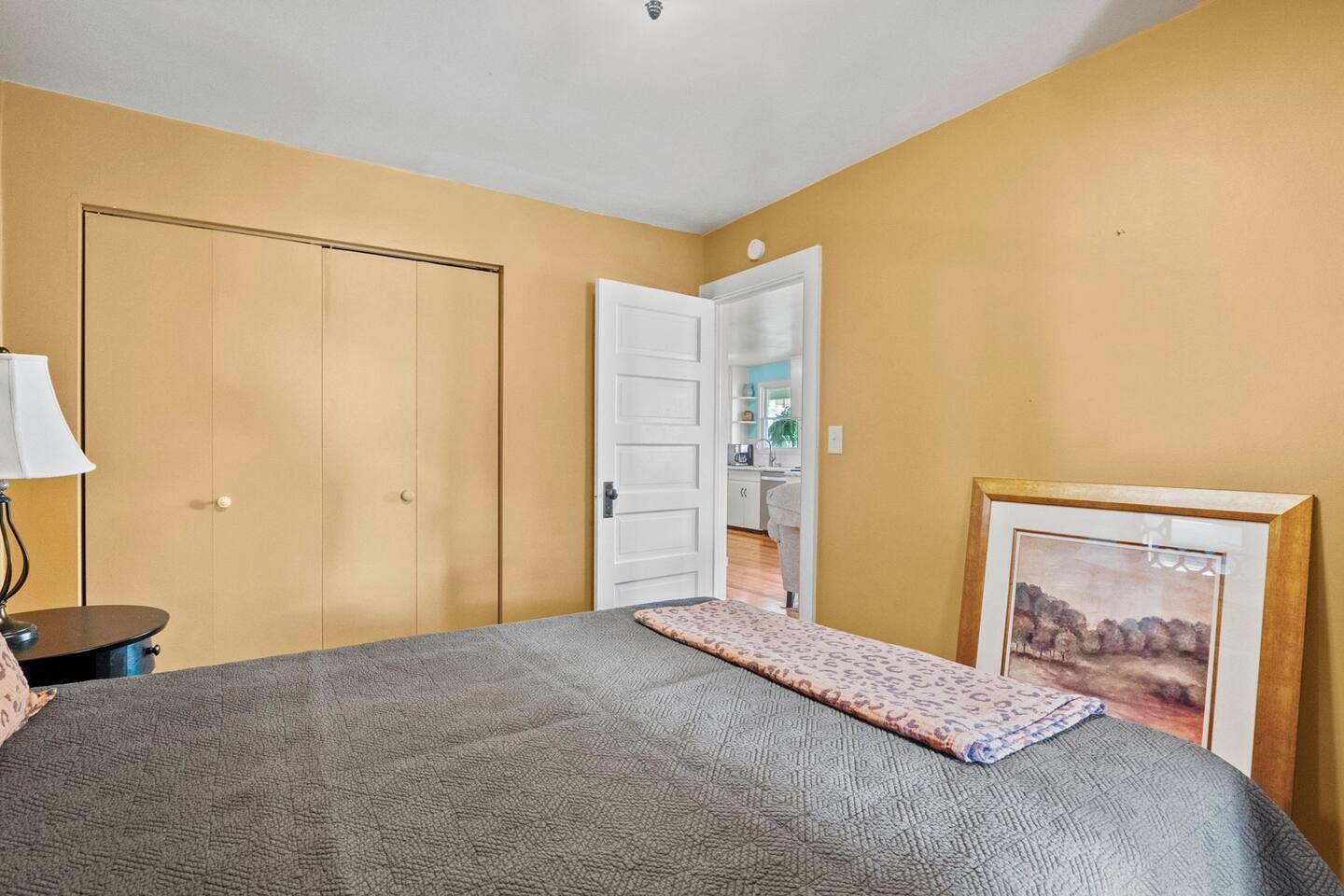 Een slaapkamer met een bed, nachtkastje, lamp, deur naar een badkamer en een lijst met een afbeelding aan de muur.