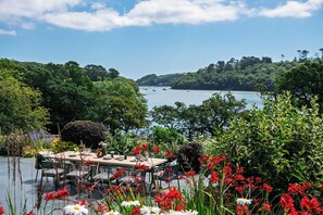 A table set outdoors with chairs, surrounded by lush greenery and a view of a lake with boats. - Calamansack Waterings - Sleeps 12 in 6 Bedrooms on the Helford River (Port Navas)
