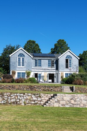 A two-story house with a stone wall in front, a solar panel on the roof, and a clear blue sky. - Calamansack Waterings - Sleeps 12 in 6 Bedrooms on the Helford River (Port Navas)