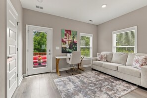 A living room with a sofa, a desk, and a chair. There are two windows with blinds, a door, and a view of greenery outside. - Stylish Antioch Stay Minutes to Downtown Nashville (Nashville)