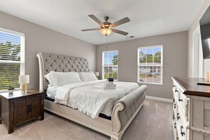 Ein Schlafzimmer mit Bett, Nachttischen, einem Schminktisch und einem Deckenventilator, sowie großen Fenstern.