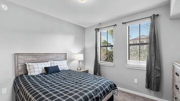 Ein Schlafzimmer mit Bett, Nachttisch, Lampe, zwei Fenstern mit Vorhängen und einer Deckenleuchte.
