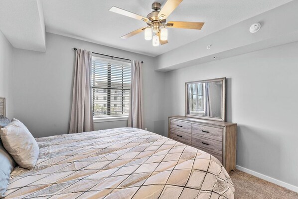Une chambre à coucher comprenant un lit, une commode, un ventilateur de plafond et une fenêtre avec des stores.