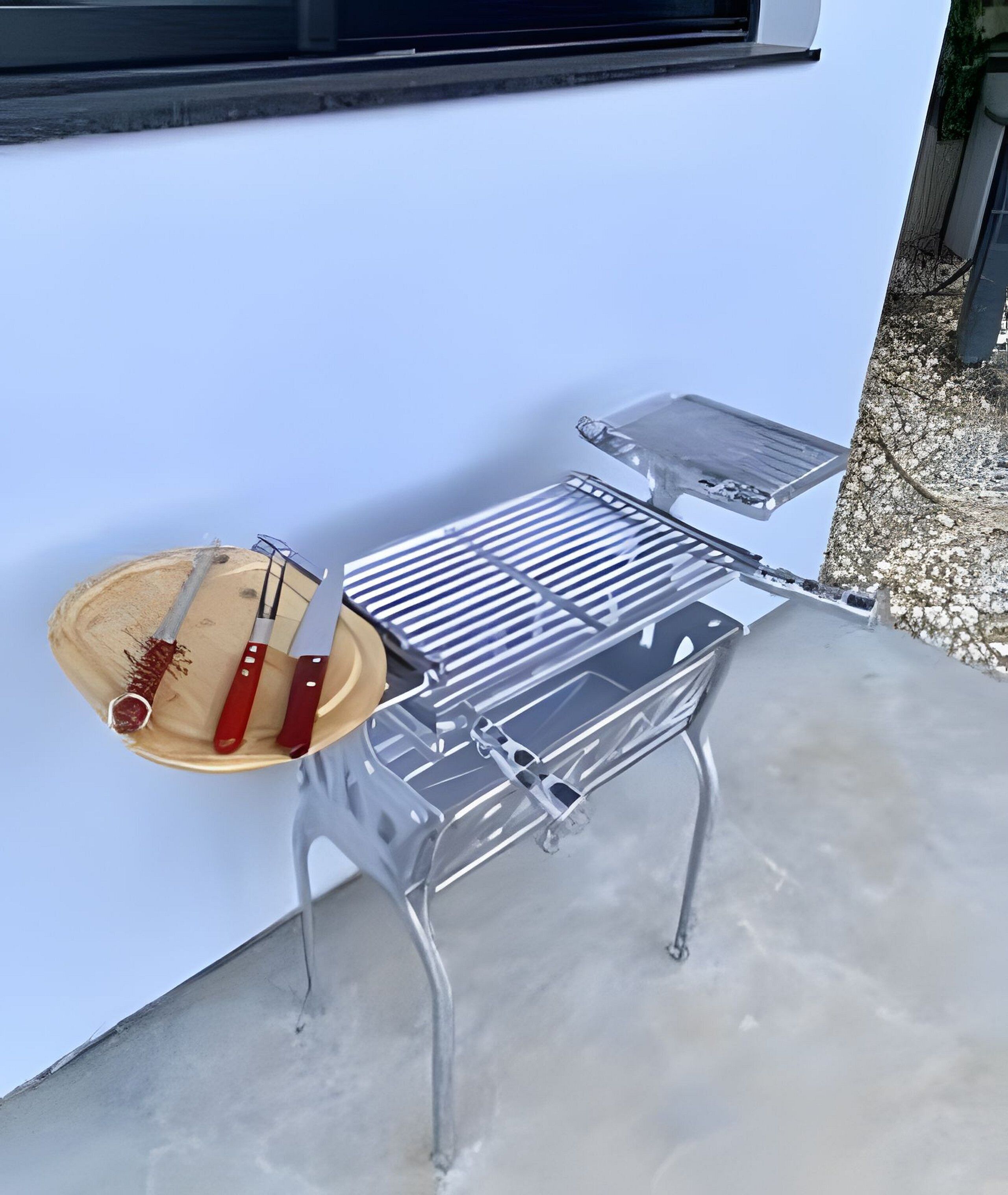 Un barbecue en métal, doté d’un plateau en bois et d’ustensiles, est posé sur une surface en béton, adossé à un mur blanc.