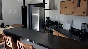 A modern kitchen with a black countertop, wooden chairs, and pendant lights. - Vista Mar Riviera - Terraço com Vista para o Mar a 50m da Praia da Ferrugem (Garopaba)