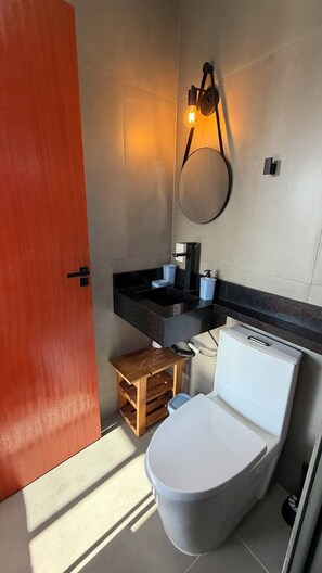 A compact bathroom with a white toilet, a wooden stool, a black countertop with a sink, and a wall-mounted light fixture. - Riviera de Garopaba  (Garopaba)