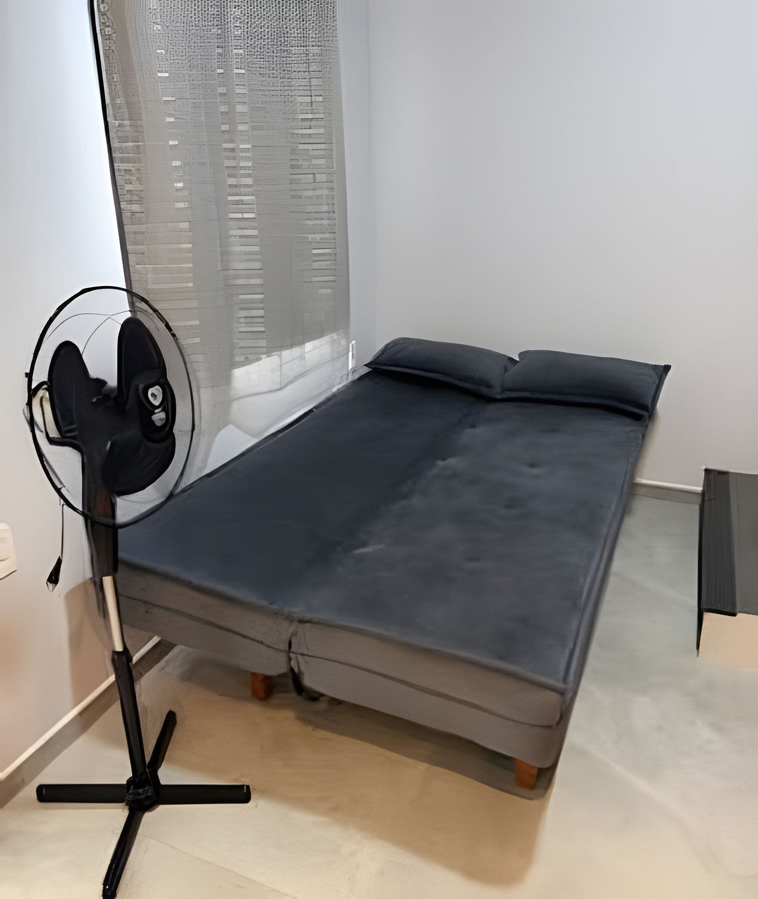 A single bed with a dark mattress and pillows, a standing fan, a window with sheer curtains, and a light-colored floor.