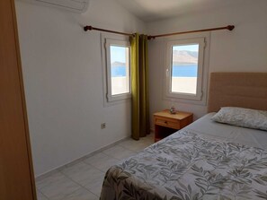 Un dormitorio con una cama, una mesita de noche, dos ventanas con cortinas, vista al mar y una montaña al fondo.