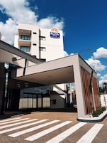 Uma entrada de hotel moderna com galeria coberta, céu claro e a placa ROTA HOTEL visível.
