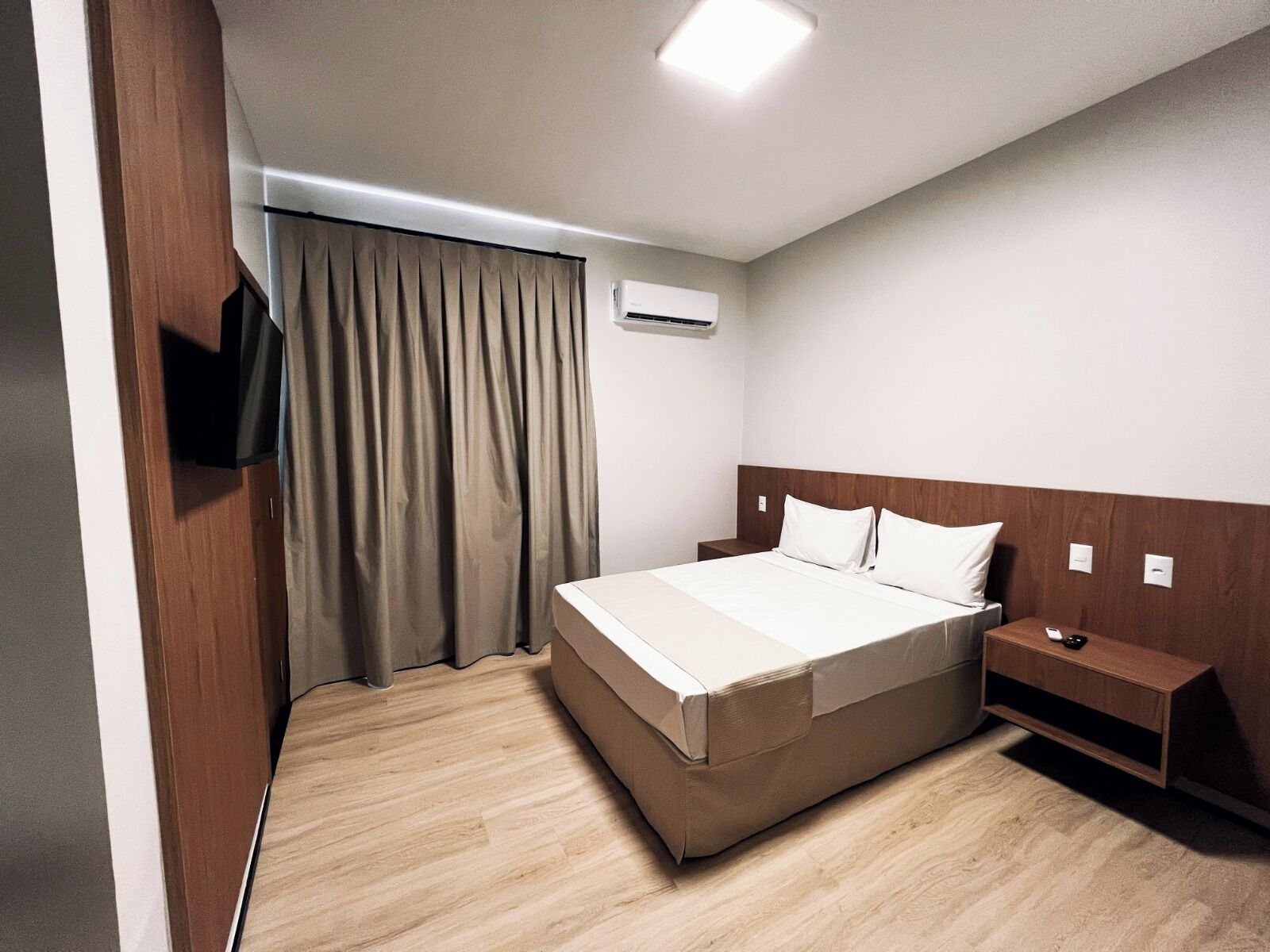 A hotel room with a bed, a nightstand, a TV, and an air conditioning unit.