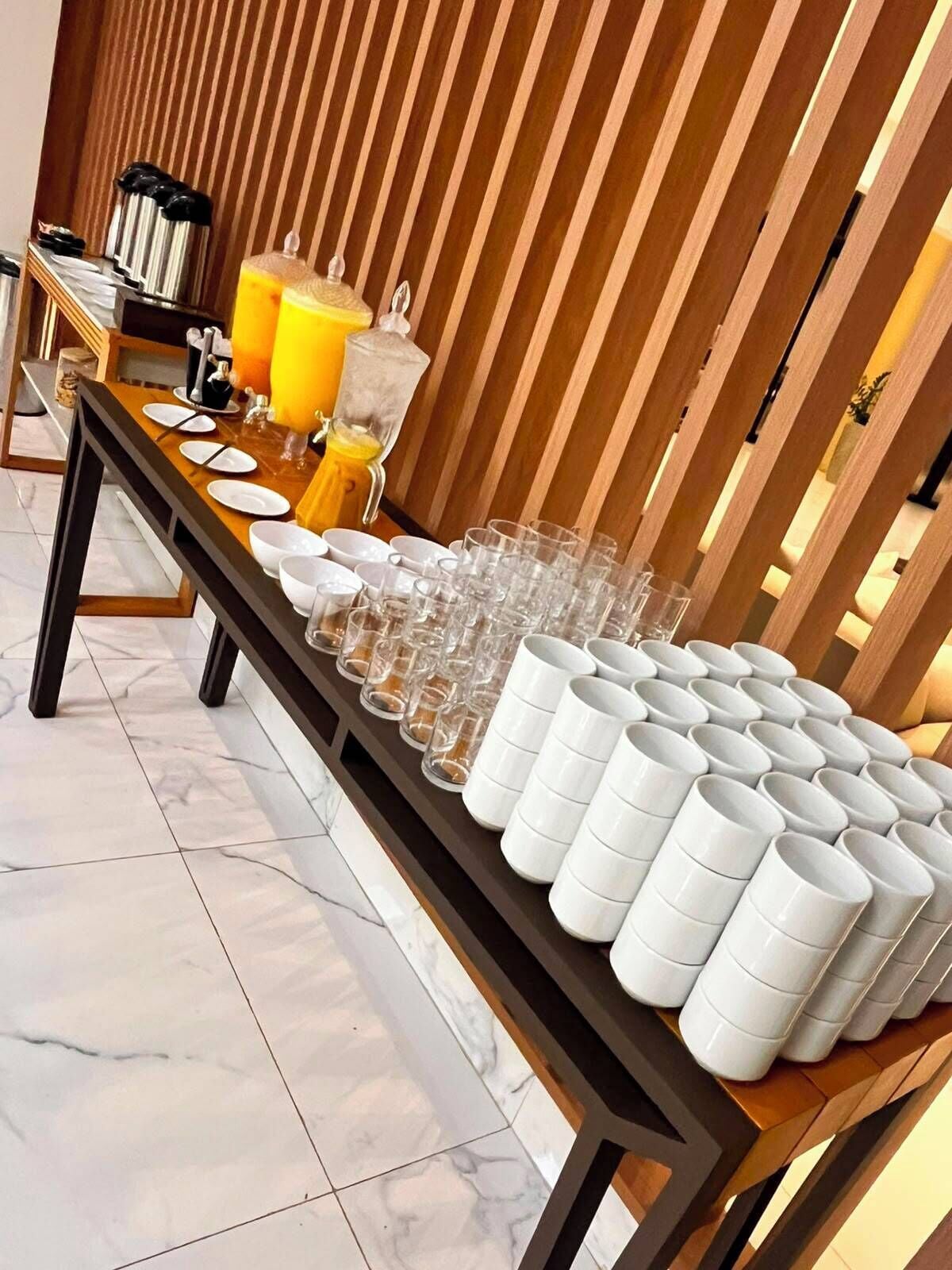 Uma mesa de buffet com diversos pratos e bebidas, uma divisória de madeira e um chão ladrilhado.