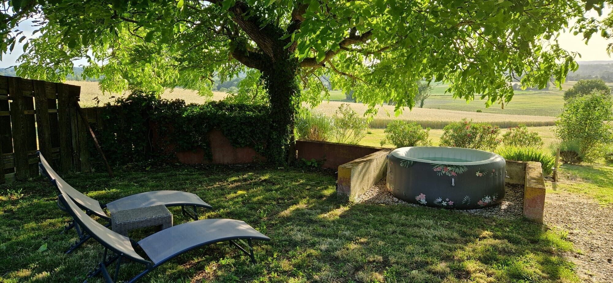 Un patio trasero con un jacuzzi, dos sillones de descanso y un árbol que da sombra.