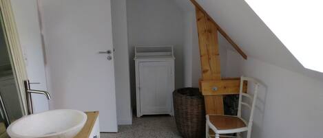 Ein kleines, weißes Badezimmer mit einer hölzernen Sitzbank, einem weißen Waschbecken und einer Tür zu einem anderen Raum.