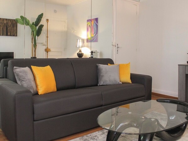 Ein Wohnzimmer mit einem grauen Sofa, zwei gelben und zwei grauen Kissen, einem Glastisch, einer Stehlampe und einem Gemälde an der Wand.